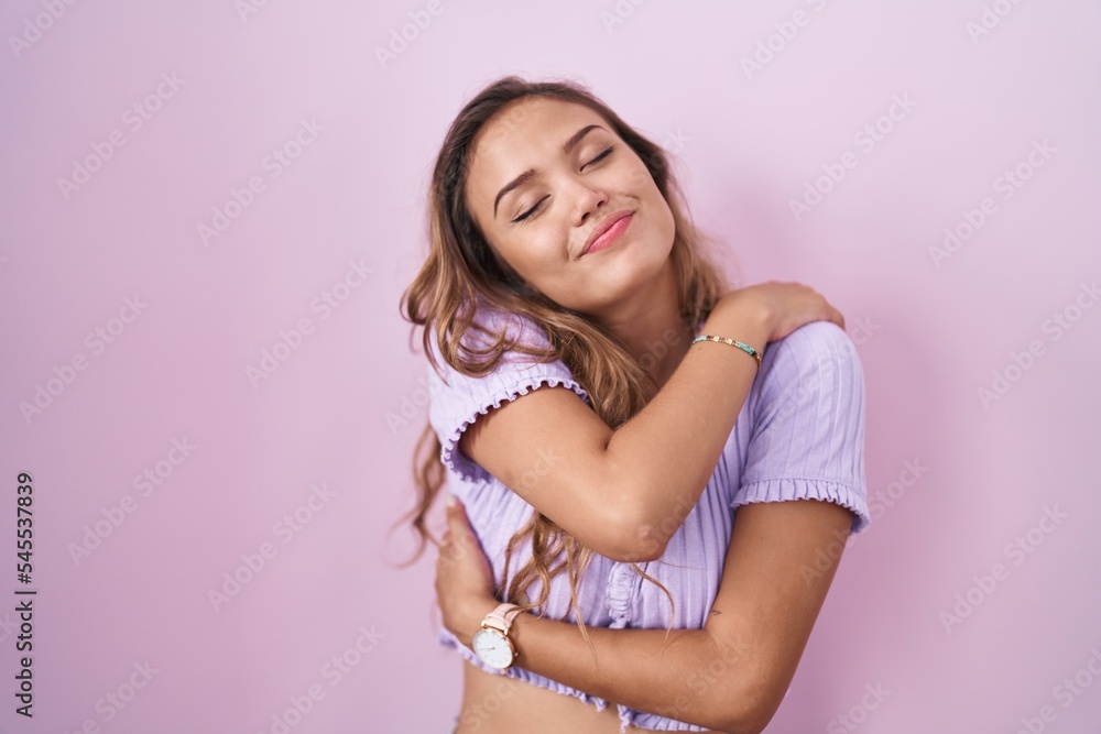 Young hispanic woman standing over pink background hugging oneself happy and positive, smiling confident. self love and self care Young hispanic woman standing over pink background hugging oneself happy and positive, smiling confident. self love and self care