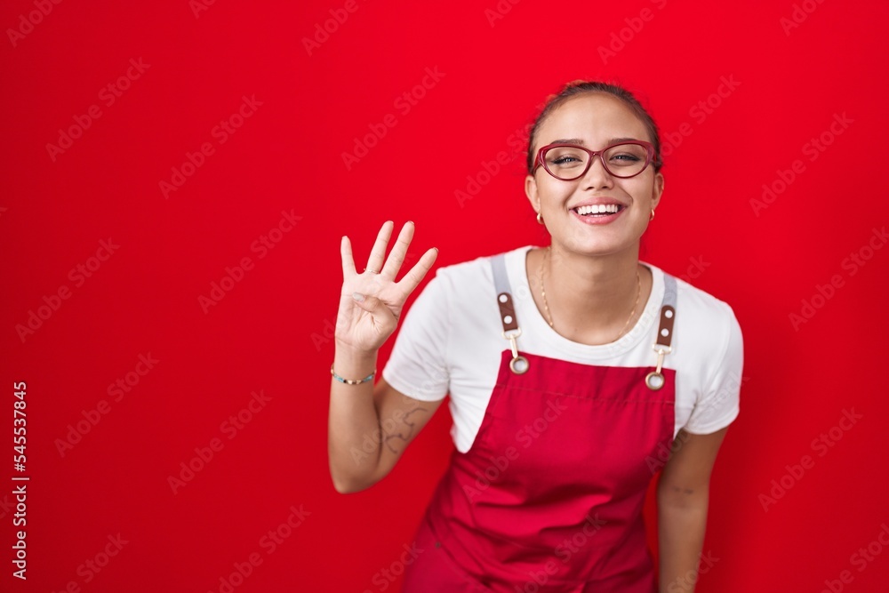 Fototapeta premium Young hispanic woman wearing waitress apron over red background showing and pointing up with fingers number four while smiling confident and happy.