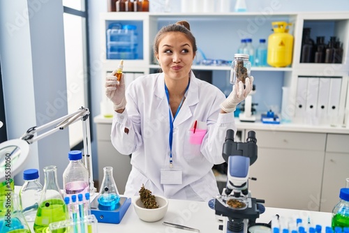 Young hispanic woman doing weed oil extraction at laboratory smiling looking to the side and staring away thinking.