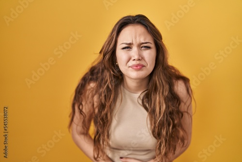 Young hispanic woman standing over yellow background with hand on stomach because indigestion, painful illness feeling unwell. ache concept.