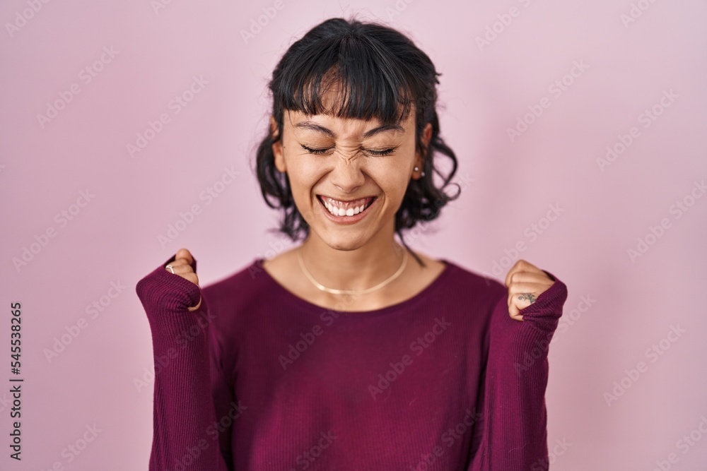 Young beautiful woman standing over pink background excited for success with arms raised and eyes closed celebrating victory smiling. winner concept. Young beautiful woman standing over pink background excited for success with arms raised and eyes closed celebrating victory smiling. winner concept.