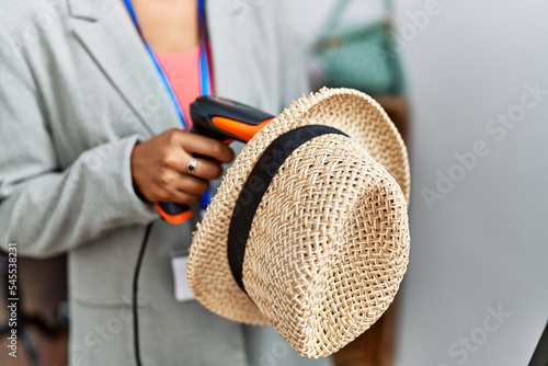 Young hispanic woman shopkeeper scanning hat using bardcode reader at clothing store photo