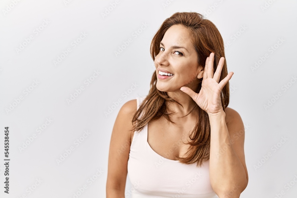 Middle age hispanic woman standing over isolated background smiling with hand over ear listening an hearing to rumor or gossip. deafness concept. Middle age hispanic woman standing over isolated background smiling with hand over ear listening an hearing to rumor or gossip. deafness concept.