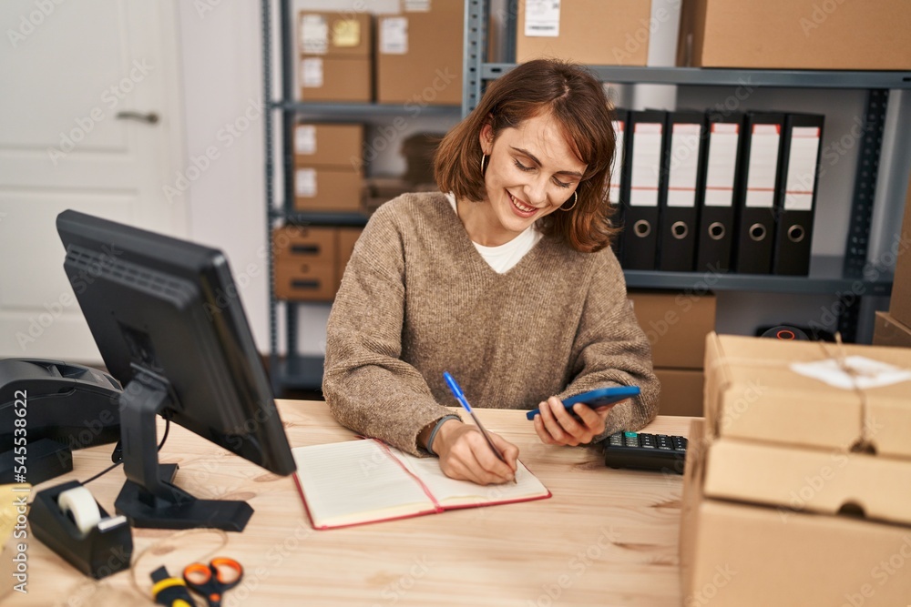 Young caucasian woman ecommerce business worker using smartphone writing on book at office Young caucasian woman ecommerce business worker using smartphone writing on book at office
