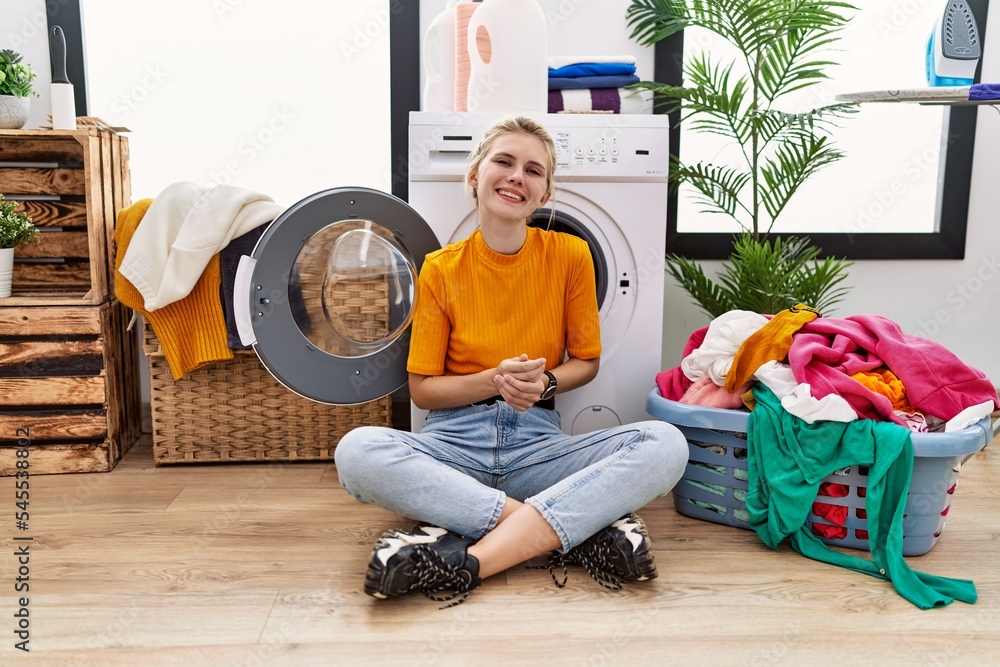 Young blonde woman doing laundry sitting by washing machine with hands together and crossed fingers smiling relaxed and cheerful. success and optimistic Young blonde woman doing laundry sitting by washing machine with hands together and crossed fingers smiling relaxed and cheerful. success and optimistic