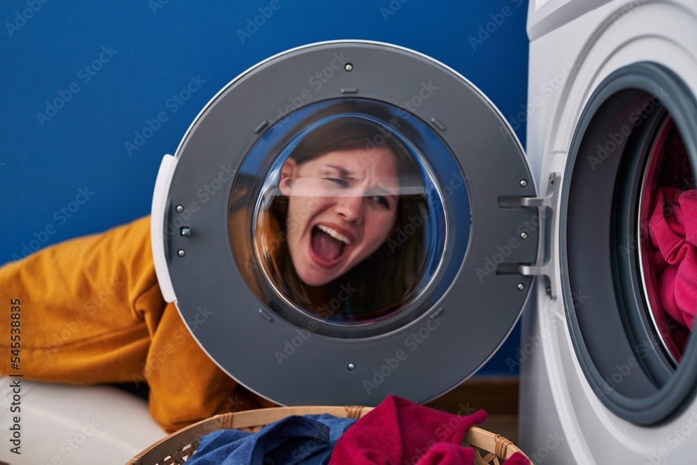 Young brunette woman looking through the washing machine window angry and mad screaming frustrated and furious, shouting with anger. rage and aggressive concept. Young brunette woman looking through the washing machine window angry and mad screaming frustrated and furious, shouting with anger. rage and aggressive concept.
