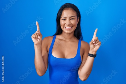 Hispanic woman standing over blue background smiling amazed and surprised and pointing up with fingers and raised arms.