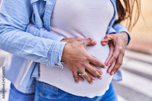 Young latin couple expecting baby touching belly hugging each other at street