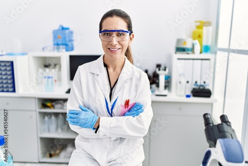 Young hispanic woman wearing scientist uniform with arms crossed gesture at laboratory