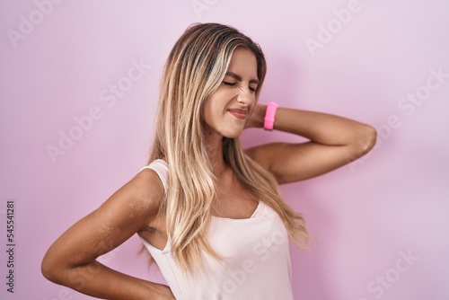 Young blonde woman standing over pink background suffering of neck ache injury, touching neck with hand, muscular pain