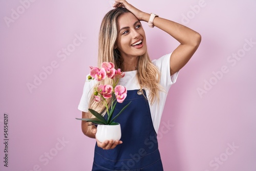 Young blonde woman wearing gardener apron holding plant smiling confident touching hair with hand up gesture, posing attractive and fashionable