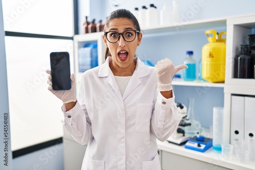 Young hispanic woman working at scientist laboratory with smartphone pointing thumb up to the side smiling happy with open mouth