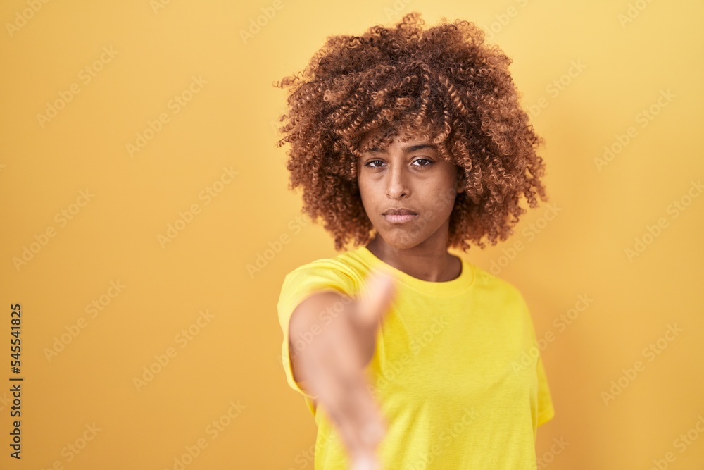 Young hispanic woman with curly hair standing over yellow background smiling friendly offering handshake as greeting and welcoming. successful business. Young hispanic woman with curly hair standing over yellow background smiling friendly offering handshake as greeting and welcoming. successful business.