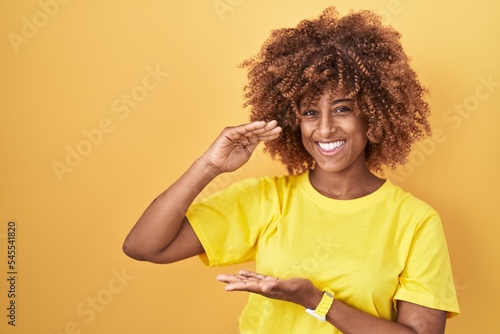Young hispanic woman with curly hair standing over yellow background gesturing with hands showing big and large size sign, measure symbol. smiling looking at the camera. measuring concept.