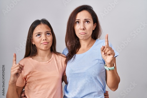 Young mother and daughter standing over white background pointing up looking sad and upset, indicating direction with fingers, unhappy and depressed.
