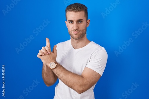 Young caucasian man standing over blue background holding symbolic gun with hand gesture, playing killing shooting weapons, angry face