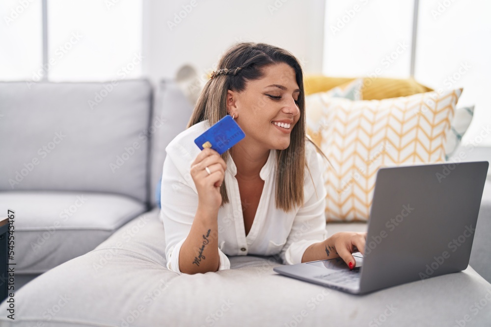 Young hispanic woman using laptop and credit card lying on sofa at home Young hispanic woman using laptop and credit card lying on sofa at home