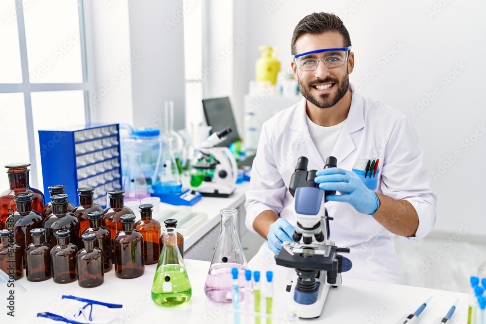 Handsome hispanic man working at scientist laboratory looking positive and happy standing and smiling with a confident smile showing teeth Handsome hispanic man working at scientist laboratory looking positive and happy standing and smiling with a confident smile showing teeth
