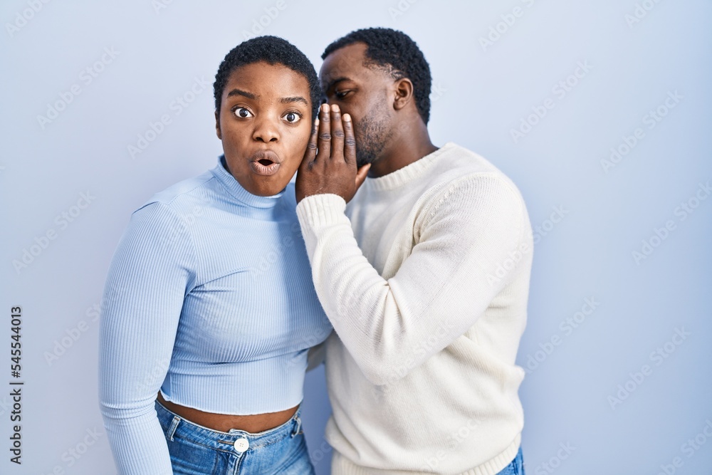 Naklejka premium Young african american couple standing over blue background hand on mouth telling secret rumor, whispering malicious talk conversation