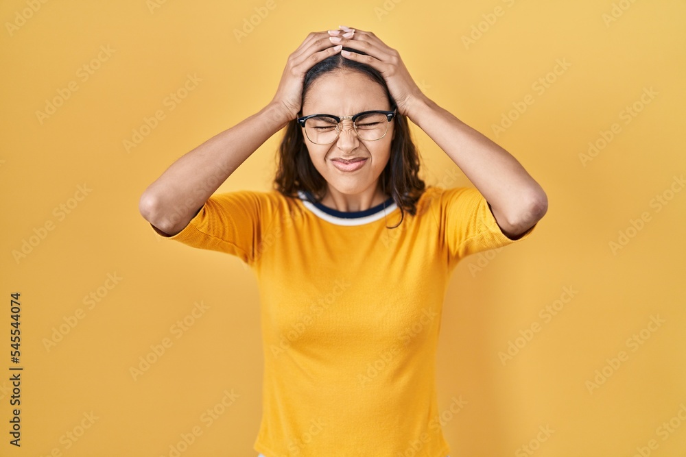 Young brazilian woman wearing glasses over yellow background suffering from headache desperate and stressed because pain and migraine. hands on head. Young brazilian woman wearing glasses over yellow background suffering from headache desperate and stressed because pain and migraine. hands on head.