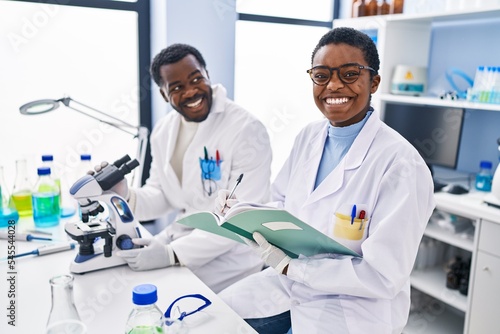 Man and woman scientists using microscope writing on notebook at laboratory