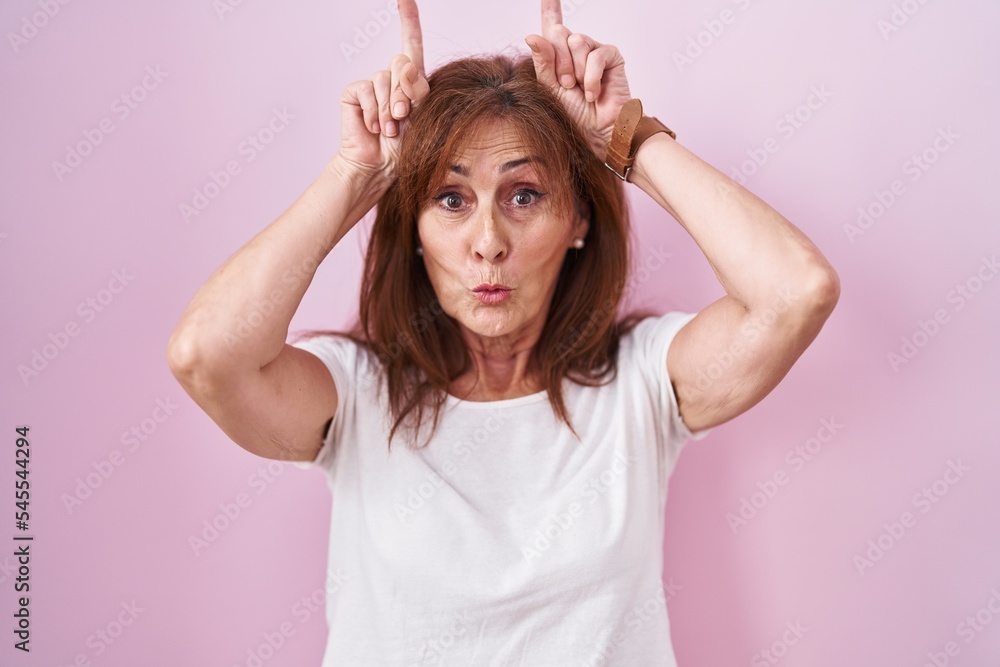 Middle age woman standing over pink background doing funny gesture with finger over head as bull horns Middle age woman standing over pink background doing funny gesture with finger over head as bull horns