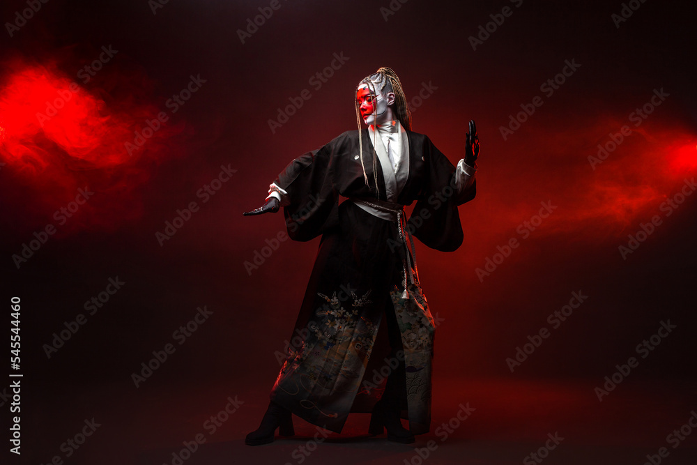 Naklejka premium A young woman in fantastic makeup and an Asian kimono, the image of a cyber geisha. Full - length photo , dancing on a dark background with red backlight