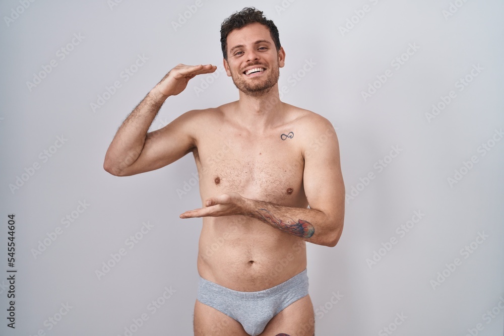 Young hispanic man standing shirtless wearing underware gesturing with hands showing big and large size sign, measure symbol. smiling looking at the camera. measuring concept.