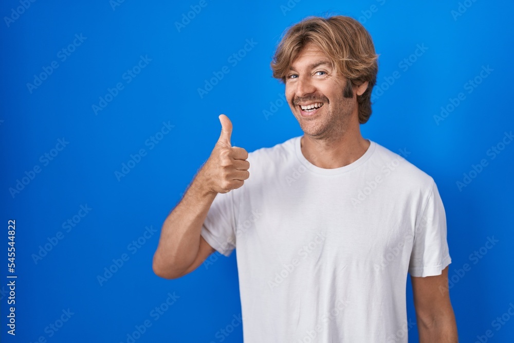 Naklejka premium Middle age man standing over blue background doing happy thumbs up gesture with hand. approving expression looking at the camera showing success.