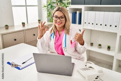 Young hispanic doctor woman holding virtual currency bitcoin pointing thumb up to the side smiling happy with open mouth