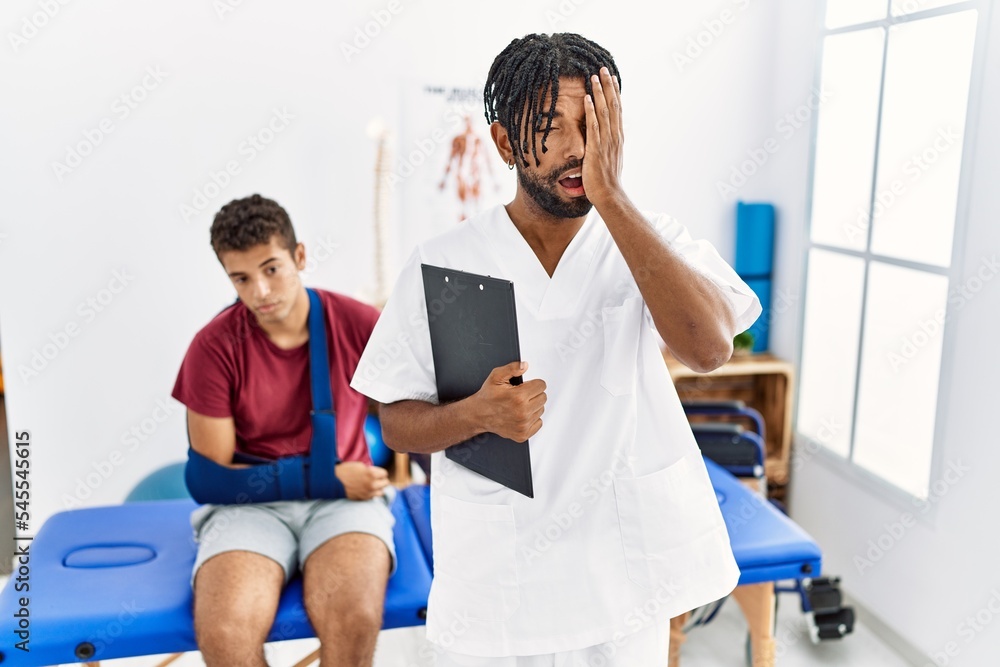 Obraz premium Young hispanic man working at pain recovery clinic with a man with broken arm yawning tired covering half face, eye and mouth with hand. face hurts in pain.