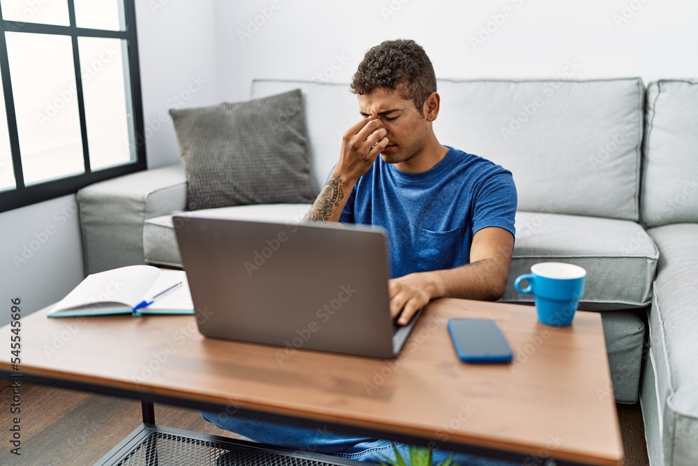 Fototapeta premium Young handsome hispanic man using laptop sitting on the floor tired rubbing nose and eyes feeling fatigue and headache. stress and frustration concept.
