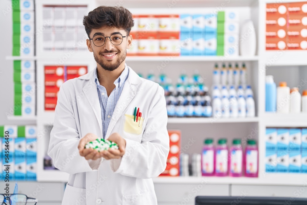 Young arab man pharmacist smiling confident holding capsules at pharmacy