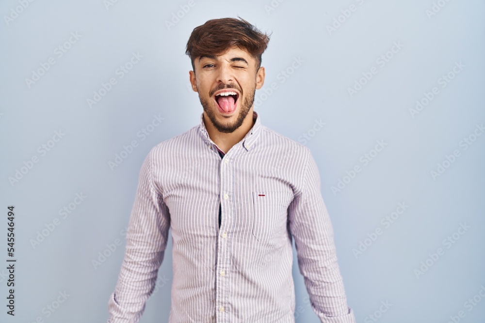 Arab man with beard standing over blue background sticking tongue out happy with funny expression. emotion concept. Arab man with beard standing over blue background sticking tongue out happy with funny expression. emotion concept.