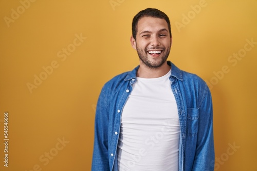 Hispanic man standing over yellow background winking looking at the camera with sexy expression, cheerful and happy face.