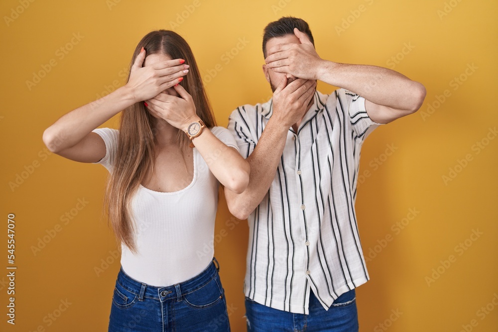 Young couple standing over yellow background covering eyes and mouth with hands, surprised and shocked. hiding emotion Young couple standing over yellow background covering eyes and mouth with hands, surprised and shocked. hiding emotion