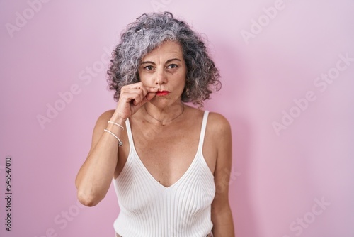 Middle age woman with grey hair standing over pink background mouth and lips shut as zip with fingers. secret and silent  taboo talking