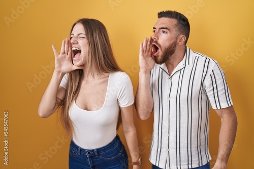 Young couple standing over yellow background shouting and screaming loud to side with hand on mouth. communication concept.