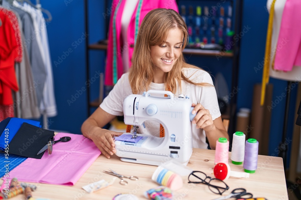 Fototapeta premium Young blonde girl tailor smiling confident using sewing machine at sewing studio