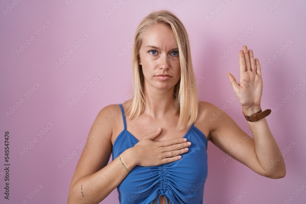 Young caucasian woman standing over pink background swearing with hand on chest and open palm, making a loyalty promise oath Young caucasian woman standing over pink background swearing with hand on chest and open palm, making a loyalty promise oath