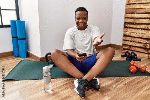 Young african man sitting on training mat at the gym using smartphone smiling cheerful offering palm hand giving assistance and acceptance.