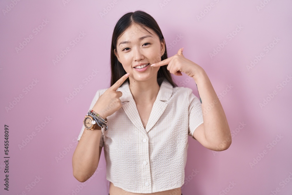 Chinese young woman standing over pink background smiling cheerful showing and pointing with fingers teeth and mouth. dental health concept. Chinese young woman standing over pink background smiling cheerful showing and pointing with fingers teeth and mouth. dental health concept.
