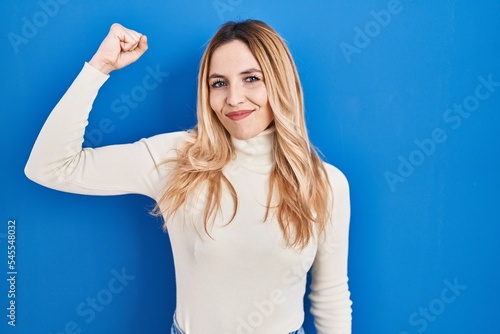 Young caucasian woman standing over blue background strong person showing arm muscle, confident and proud of power