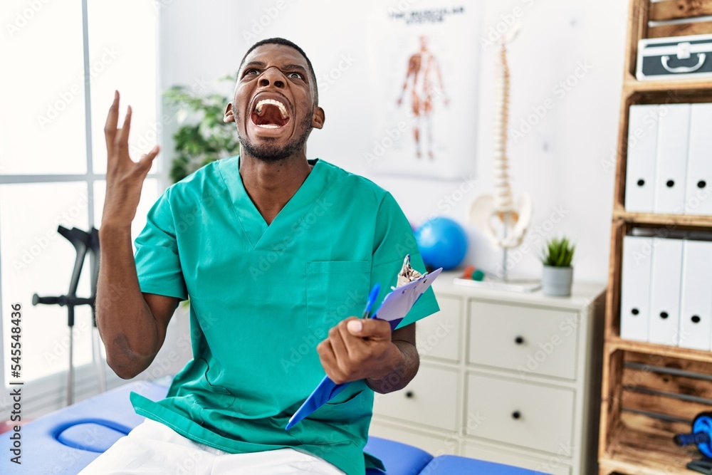 Fototapeta premium Young african american man working at pain recovery clinic crazy and mad shouting and yelling with aggressive expression and arms raised. frustration concept.