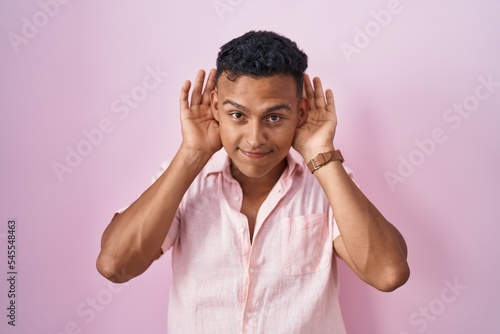 Young hispanic man standing over pink background trying to hear both hands on ear gesture, curious for gossip. hearing problem, deaf