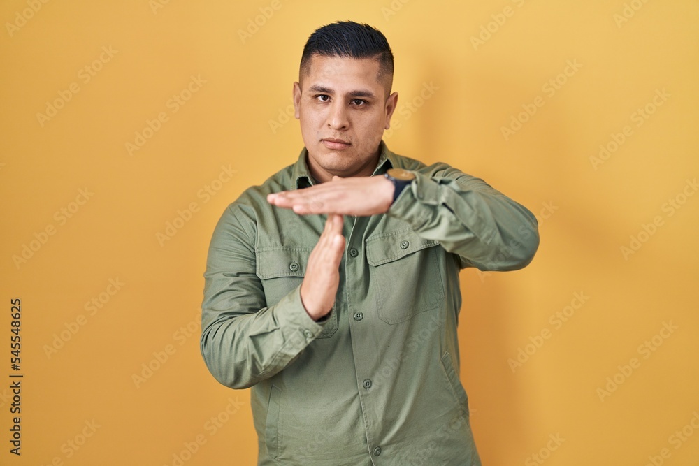 Hispanic young man standing over yellow background doing time out gesture with hands, frustrated and serious face Hispanic young man standing over yellow background doing time out gesture with hands, frustrated and serious face