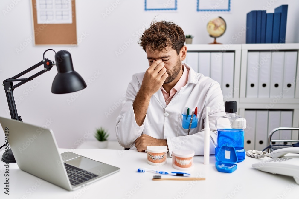 Young hispanic dentist man working at medical clinic tired rubbing nose and eyes feeling fatigue and headache. stress and frustration concept. Young hispanic dentist man working at medical clinic tired rubbing nose and eyes feeling fatigue and headache. stress and frustration concept.