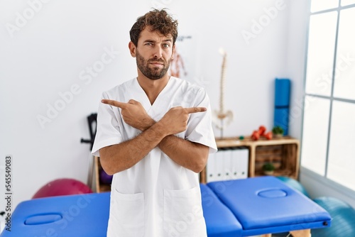 Young handsome physiotherapist man working at pain recovery clinic pointing to both sides with fingers  different direction disagree