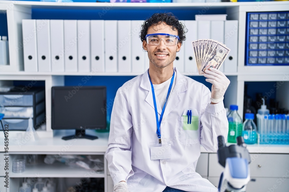 Hispanic man working at scientist laboratory holding money looking positive and happy standing and smiling with a confident smile showing teeth Hispanic man working at scientist laboratory holding money looking positive and happy standing and smiling with a confident smile showing teeth
