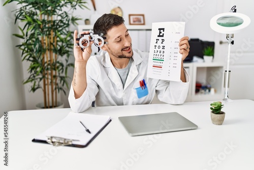 Young hispanic man wearing optician uniform holding snellen test at clinic photo